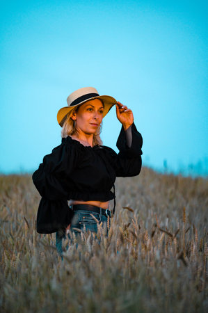 A blonde woman in a black top and jeans stands in a field of wheat at sunset, holding her hat. The sky is a clear blue.の写真素材