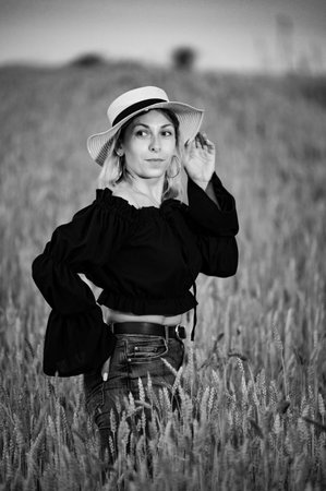 A black and white portrait of a woman with blonde hair, wearing a straw hat and a black top, in a field of tall wheat. She's captured in a serene moment, surrounded by the natural beauty of the field.の写真素材