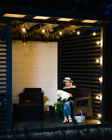 A woman in a hat sits in a wicker chair on a patio, illuminated by string lights. She's carefully arranging a bouquet of white flowers. The scene is peaceful and serene.の写真素材