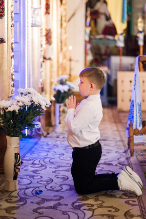 A young boy, dressed in formal attire, kneels in prayer before a religious icon inside a church. White flowers are visible in a vase nearby.の写真素材