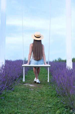 Woman in straw hat sits on a swing amidst a lavender field, enjoying a peaceful moment.の写真素材