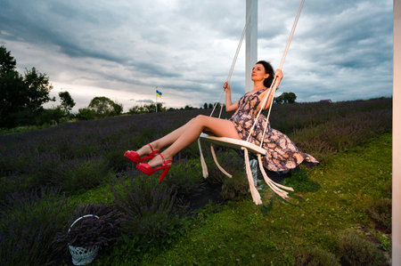 A woman in a floral dress enjoys a swing ride in a vibrant lavender field. The sky is cloudy, creating a serene atmosphere.の写真素材