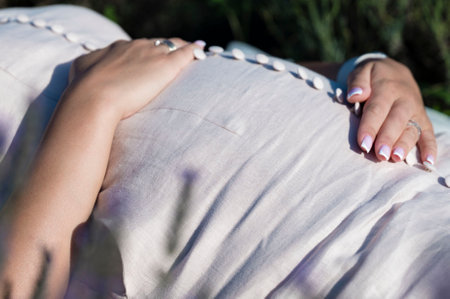 Close-up of a pregnant woman's belly, hands resting gently. She's wearing a light-colored dress and is outdoors in a sunny, peaceful setting.の写真素材