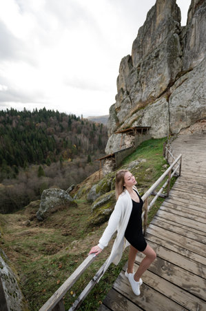 A young woman wearing a white cardigan and black dress stands on a wooden walkway overlooking a mountainous landscape. The scene is serene and peaceful.の写真素材