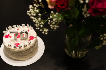A delicious-looking white cake, decorated with red accents and silver '70' candles, sits on a table next to a bouquet of red roses. A celebratory scene.の写真素材