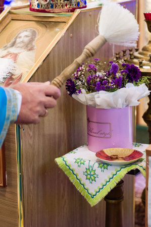 A hand holds a ceremonial brush near a floral arrangement on a small table. Religious icon and gold accents are visible. The scene suggests a sacred ritual.の写真素材