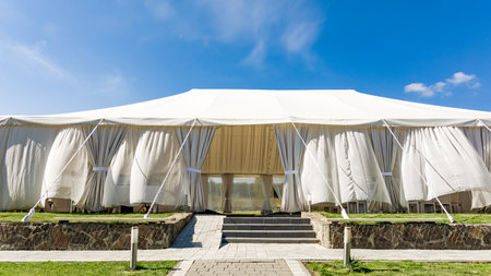 A spacious white event tent stands on a grassy lawn under a bright blue sky. Curtains adorn its sides, and stone steps lead to its entrance. Ideal for outdoor gatherings.の写真素材