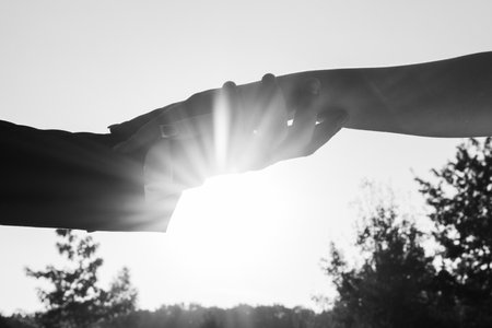 A black and white image of two hands meeting, silhouetted against a sunlit sky. The scene evokes feelings of connection and hope.の写真素材