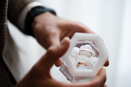 A person's hands gently hold a modern, white hexagonal box containing gold wedding bands. The image evokes feelings of anticipation and celebration.の写真素材