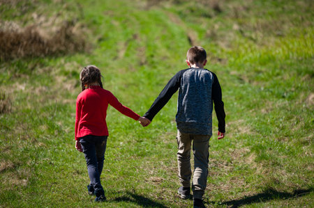 Two young siblings, a boy and a girl, walk hand-in-hand along a verdant path. The sun shines...
