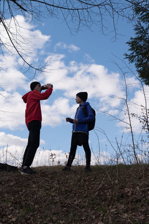 Silhouetted against a vibrant blue sky, two young people pause on a hill during a hike. One drinks from a container, the other looks on. Dry grass and leaf litter cover the ground.の写真素材
