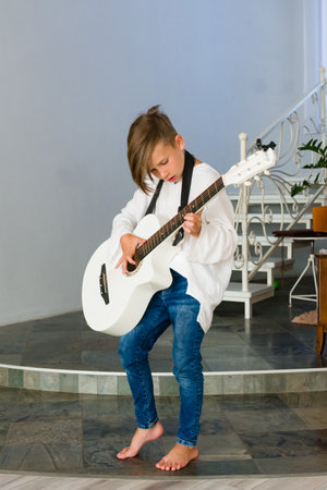 A child with shoulder-length hair plays a white acoustic guitar in a home setting. They are barefoot, wearing jeans and a white shirt, focused on their music.の写真素材