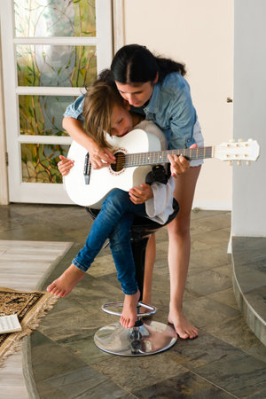 A woman gently guides her child's hands as they learn to play a white acoustic guitar. The setting is a home, intimate and warm.の写真素材