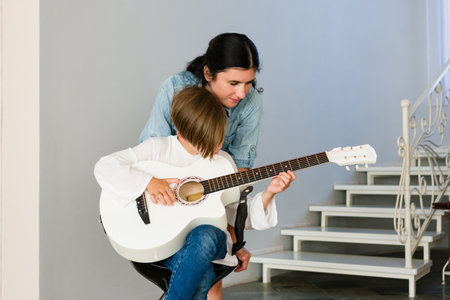 A young child sits on a stool, receiving a guitar lesson from an adult. They focus intently on the...