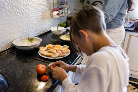 A child meticulously cuts tomatoes to prepare appetizers. The kitchen is modern, with a dark countertop and white tiled backsplash. A plate of prepared snacks sits nearby.の写真素材