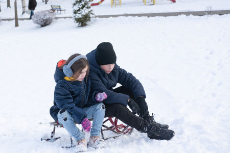 Two children, a boy and a girl, sit on a sled in the snow. They are bundled up in warm winter...