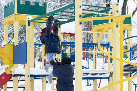 A child climbs a colorful playground structure covered in snow. An adult assists the child. Winter wonderland setting.の写真素材