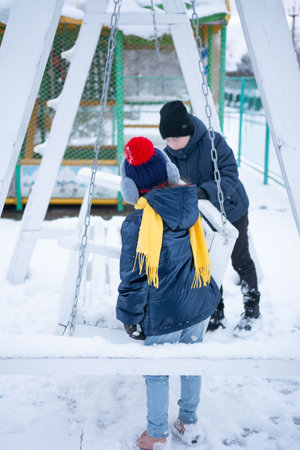 Two children, wearing winter hats, scarves, and coats, are sitting on a snow-covered swing set. The swing set is made of white wood and is located outdoors in a snowy area.の写真素材