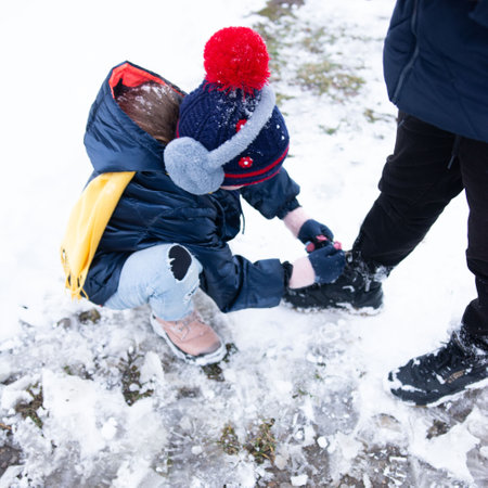 A young child, wearing a warm hat and jacket, assists an adult by fastening their winter boots. The ground is covered in snow and ice. A heartwarming scene of winter assistance.の写真素材