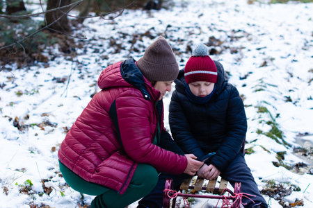 An adult and a young child sit on a sled in a snowy forest, looking down at a mobile phone. They are bundled in warm winter clothing.の写真素材