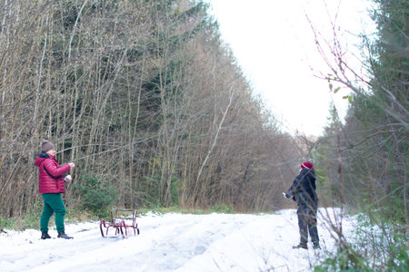 A winter scene depicts two people engaging in a snowball fight along a snowy path through a leafless forest. A small sled sits unused nearby.の写真素材