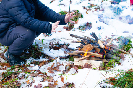 A person crouches near a small bonfire built on the snowy ground. Dry twigs and branches burn...