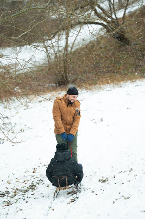 A joyful father, wearing a brown jacket, pulls his child on a small sled through a snowy wooded...