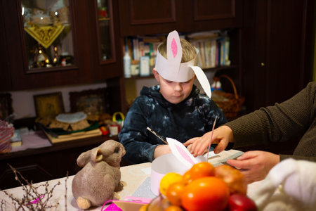 A young child wearing a bunny ear headband sits at a table, carefully crafting a paper rabbit mask. An adult assists, creating a heartwarming Easter scene.の写真素材
