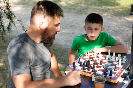 A man and a boy are concentrating on a chess match. They're seated at a wooden table in a park-like...