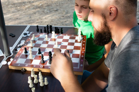 Focused on a chessboard, an adult and a child engage in a game of chess. The setting is outdoors, likely a park or recreational area.の写真素材
