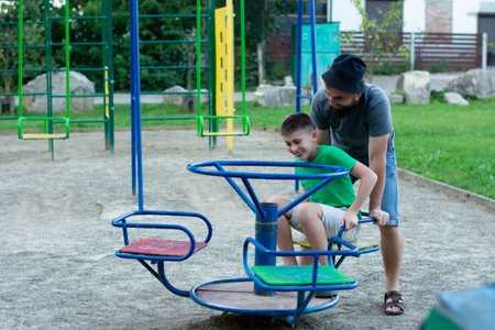 A father gently assists his son as they spin on a small merry-go-round at a playground. The scene is cheerful and depicts a loving father-son bond.の写真素材