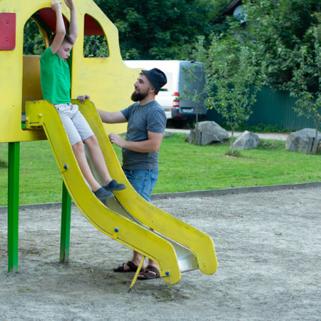 A bearded man helps a boy onto a yellow playground slide in a park setting. The boy is wearing a green shirt and shorts. It's a sunny day. They appear happy and enjoying their time together.の写真素材