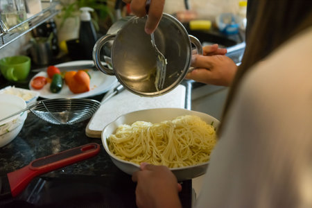 In a kitchen setting, a person is adding cooked pasta from a pot into a white bowl. Other food prep items are visible on the counter.の写真素材