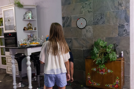 A young girl with long hair aims at a dartboard mounted on a stone wall in a home kitchen. An adult partially visible behind her. The kitchen features a modern island and traditional decor.の写真素材