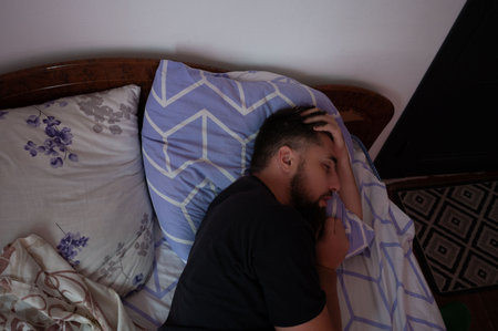 A man with a beard is lying in bed with patterned pillows and bedding, eyes closed, and his hand...