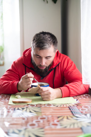 Indoor shot of a bearded man in a red jacket, focusing intently on his soup, with bread and a patterned table adding warmth. Natural light filters in, creating a comforting, personal scene.の写真素材