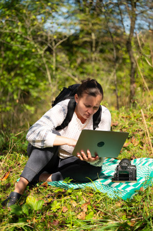 A woman sits on a patterned blanket outdoors, using her laptop. She is wearing a sweater, pants, and a backpack. A camera sits beside her on the blanket.の写真素材