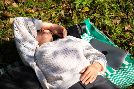 A woman is relaxing on a blanket in nature with a laptop, phone, and camera beside her. She is...