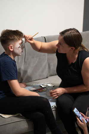 Indoor scene: A woman carefully applies white face paint to a boy's face using a brush. They sit on a couch, creating a relaxed and intimate atmosphere.の写真素材
