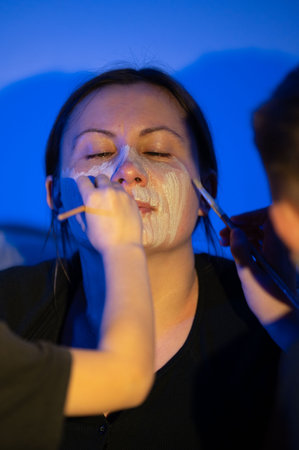 Close-up view of a person having a white facial mask applied with brushes. The setting appears to be indoors, under blue lighting. The scene is intimate and focused on the application process.の写真素材