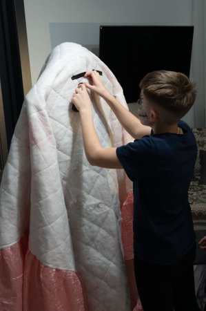 A young person with face paint carefully applies black tape to a white quilted fabric ghost costume, creating eyes and a mouth. The setting appears to be a home, possibly during Halloween preparationsの写真素材