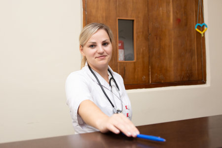 A kind female doctor, with blonde hair and a stethoscope around her neck, extends a pen across a brown desk. She's wearing a white coat, ready to assist patients. Wooden background.の写真素材
