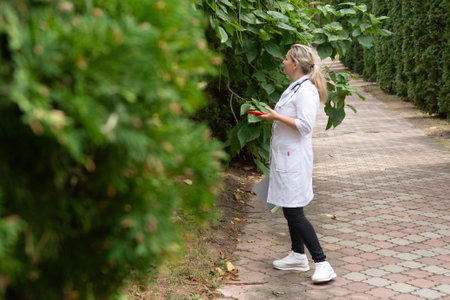 A healthcare professional wearing a white lab coat and holding a notepad stands next to green foliage along a pathway. The setting is outdoors with trees lining the path on a bright day.の写真素材