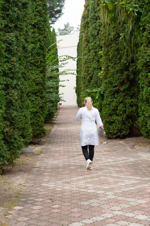 A woman in a white coat walks down a brick pathway, flanked by towering green evergreen trees. The scene evokes tranquility in this natural setting. The path leads towards a building.の写真素材