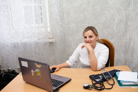 A medical professional in a white lab coat is seated at a desk, working on a laptop and handling a mouse. A sphygmomanometer sits on a table in a office setting. The wallpaper is neutral.の写真素材