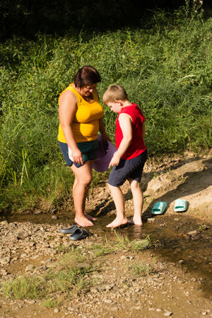 A woman and a boy are standing in a shallow stream on a sunny day. They are looking down at something in the water. The surrounding area is grassy and rural.の写真素材