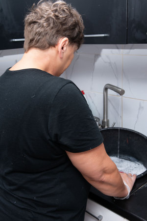 An older woman in a black top stands at a kitchen sink, wearing gloves and washing it with water running from the faucet. The scene captures domestic life, cleanliness, and daily routines.の写真素材