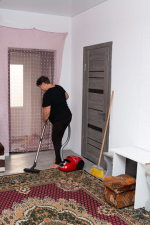 An interior image shows a woman in a black T-shirt vacuuming a patterned carpet in a room with a door, white walls, and a stool. Natural light streams in, enhancing the tidy atmosphere.の写真素材