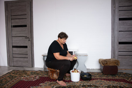Middle-aged woman is sitting on a stool peeling potatoes next to a white bucket inside a room with two doors. The scene is set on an ornamental carpet, evoking a domestic and traditional feel.の写真素材