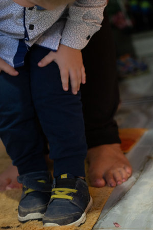 The photo shows a child wearing dark blue trousers and shoes with yellow laces, standing near an adult's feet. The photo gives a close-up view of the child's outfit.の写真素材